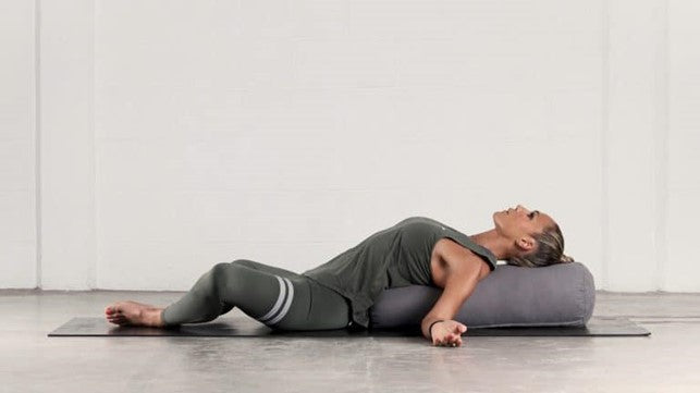 Woman laying on a black yoga mat with a pillow propped under her back for support.