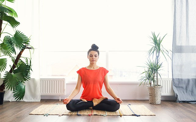 Woman seated practicing yoga/meditating on the floor in front of a window and two plants.